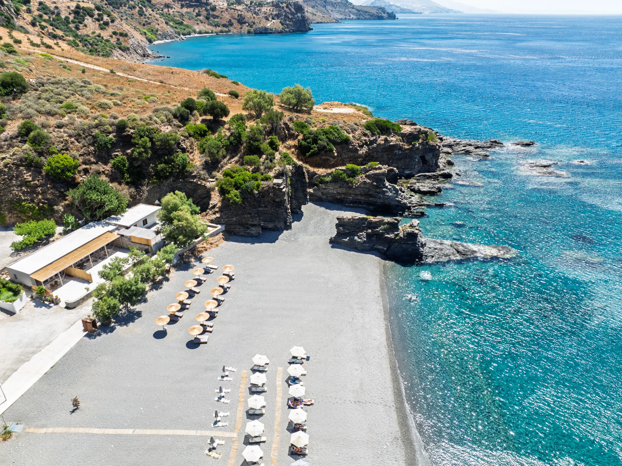 High over Korakas — the umbrella rows on the grey pebbles, the rocky point, and the coastline of south Crete running east