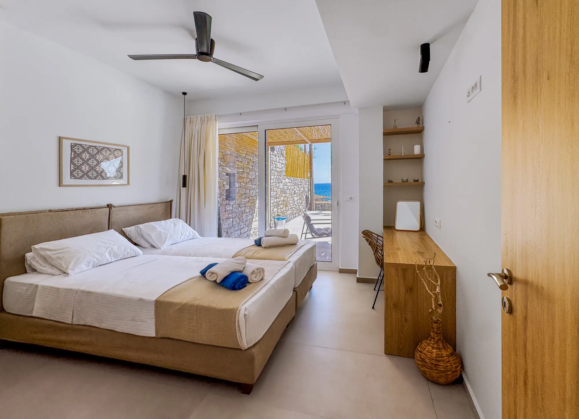 A bedroom with two single beds, folded towels, a desk in oak, and the glass door opening to the stone wall of the terrace and the sea
