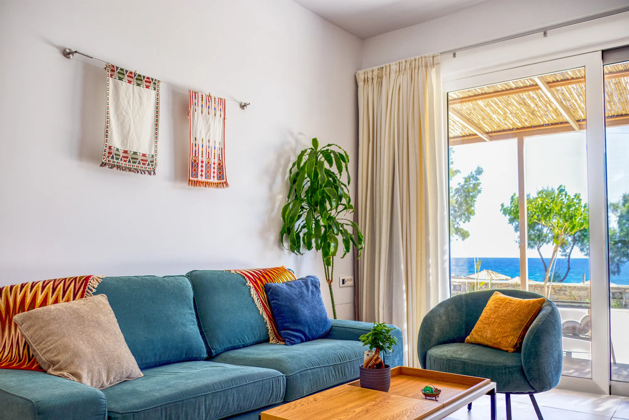 The living room — teal sofa with woven Cretan textiles on the wall, sheer curtains, and the sea through the open terrace door