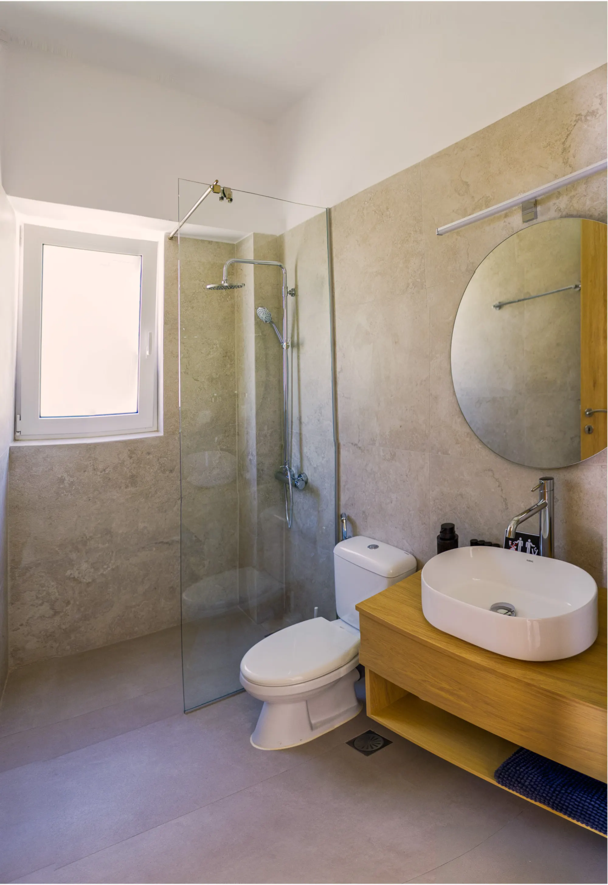 The bathroom — walk-in rain shower behind glass, stone-finished walls, round mirror and white basin on an oak vanity
