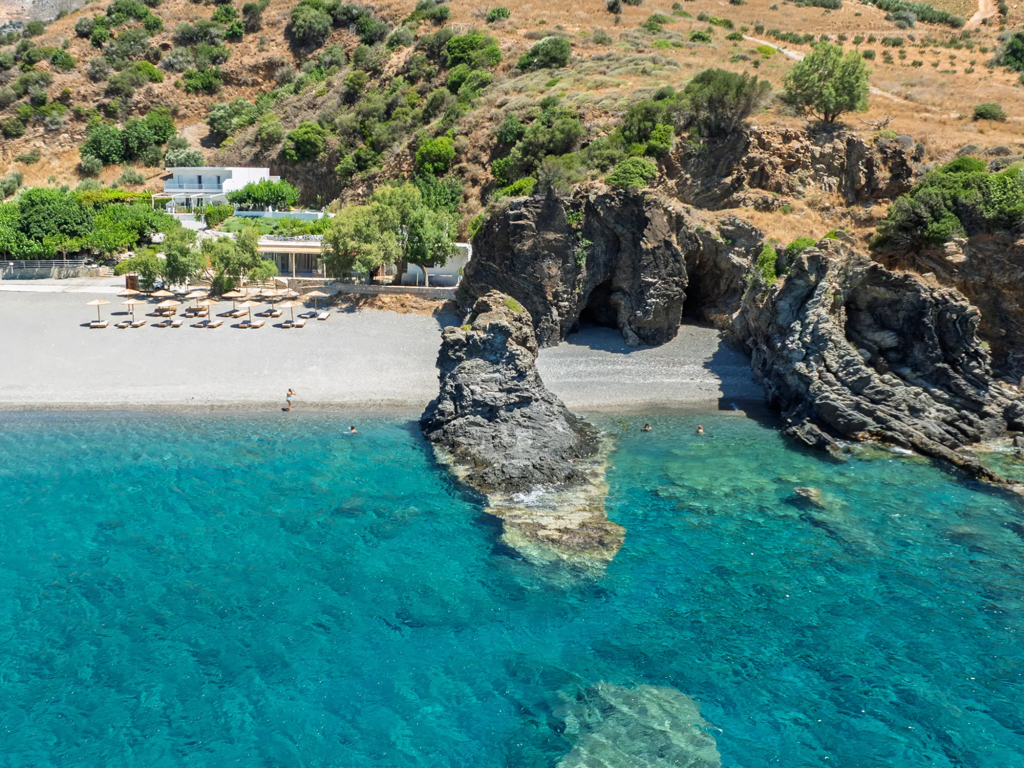 The sea caves and rock arch of Korakas seen from over the water — swimmers below the cliffs, the house among the greenery behind the beach