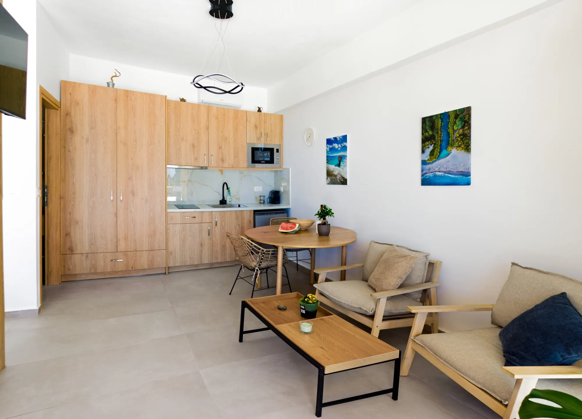 The studio's sitting corner — pale wood kitchenette, round dining table, two cushioned armchairs under prints of the coast