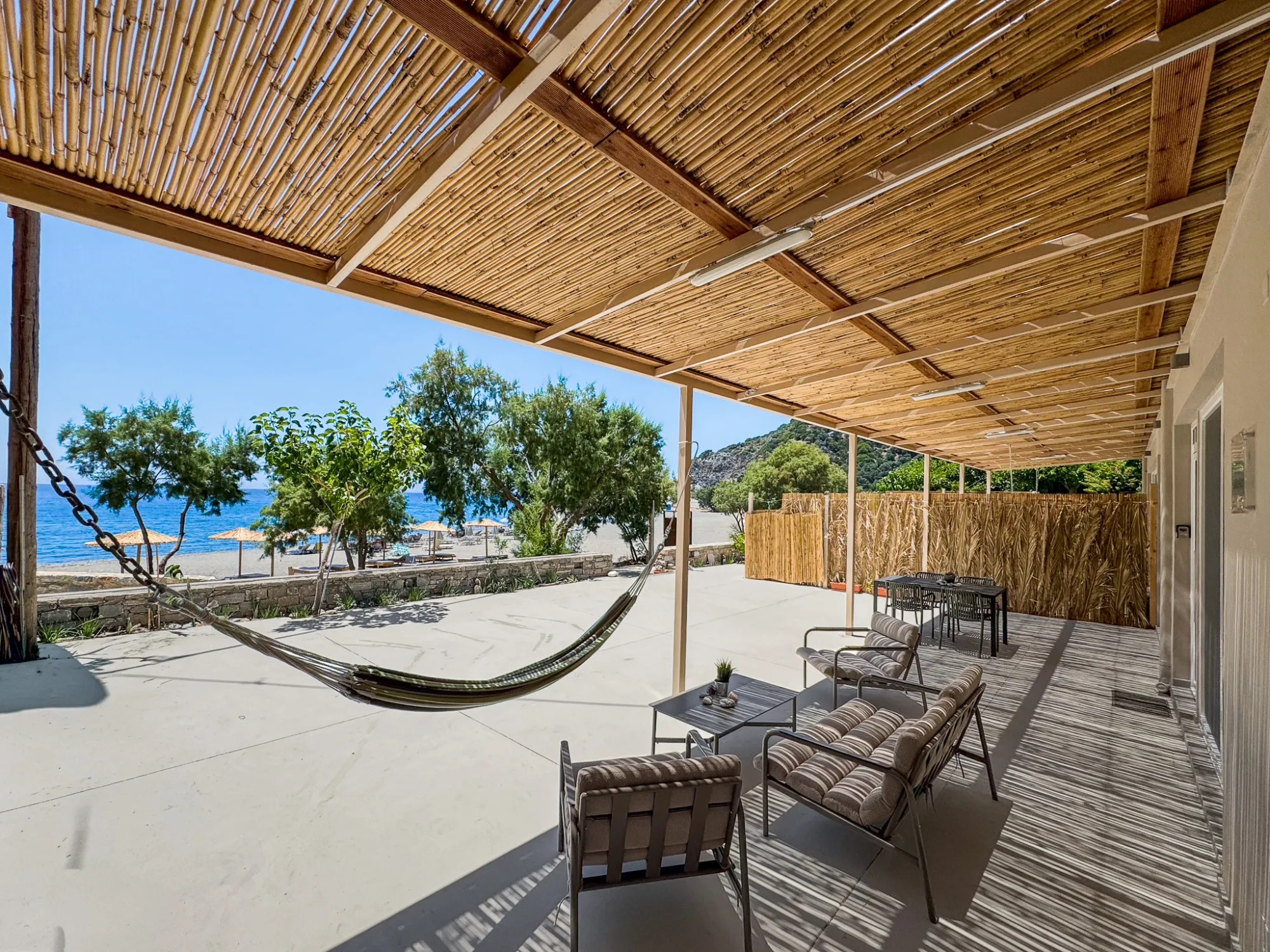 Under the bamboo pergola on the terrace — the hammock strung between posts, lounge seating and dining table, the low stone wall and the beach and sea beyond